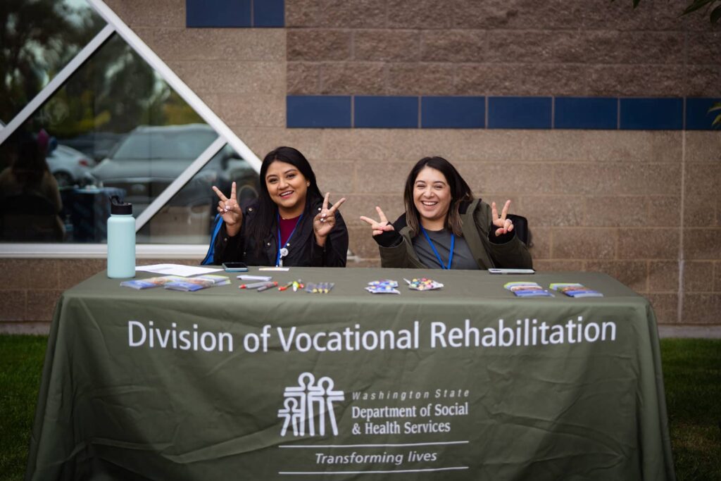 Two women are sitting behind a table that has a khaki colored tablecloth with the words Department of Vocational Rehabilitation; they are each holding up peace signs on each hand.