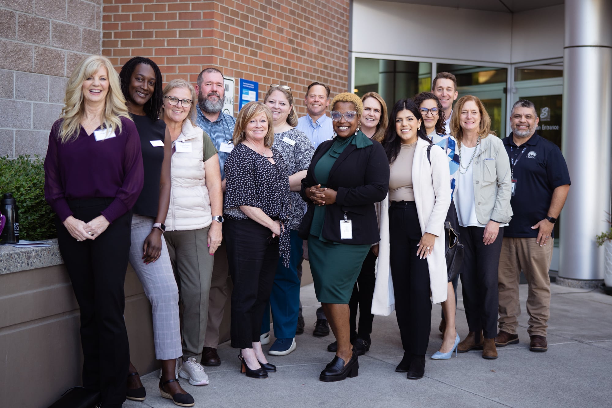 a diverse group of 13 people in professional attire stand together outside the entrance to a brick hospital