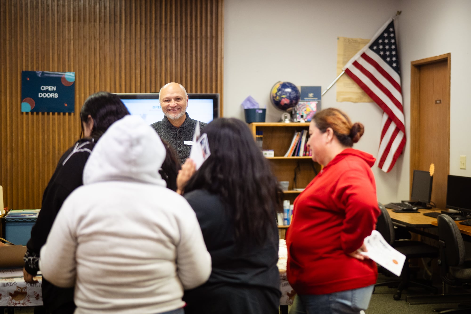 One light skinned smiling man faces toward the camera and is surrounded by a group of people with their backs toward the camera as they listen to him talk in a classroom.