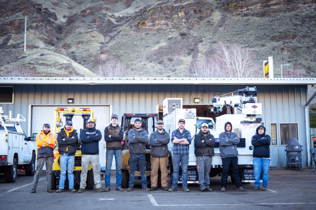 A team of ten irrigation technicians lined up elbow to elbow with their arms cross in front of their chests, the company building in the background and a giant rocky hillside in the distance.