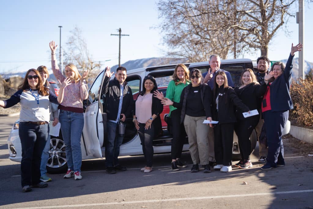 A diverse group of community nonprofit representatives enthusiastically smile and wave outside a white minivan beneath a blue sky.