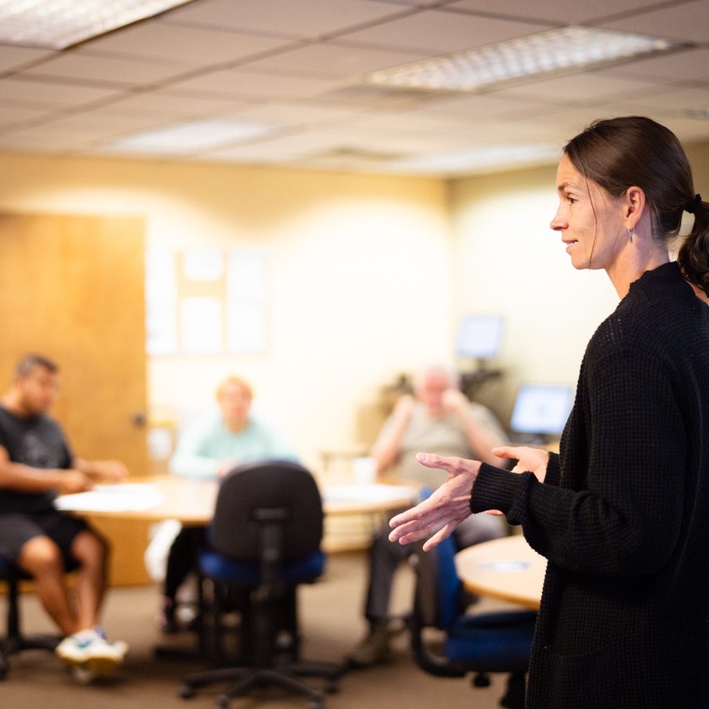 A woman with a ponytail wearing a black blazer stands at the front of a workshop room where people are seated in the background around tables.