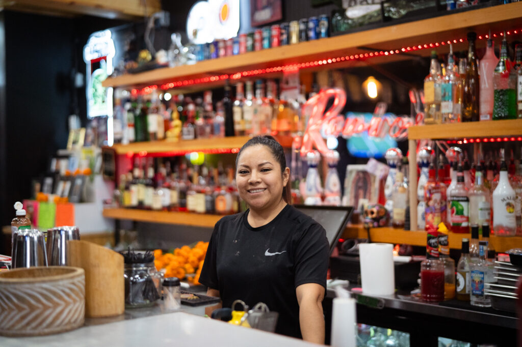 10-30-24 Ramiro’s-4 Woman with brunette hair in a ponytail standing behind a table while smiling at the camera