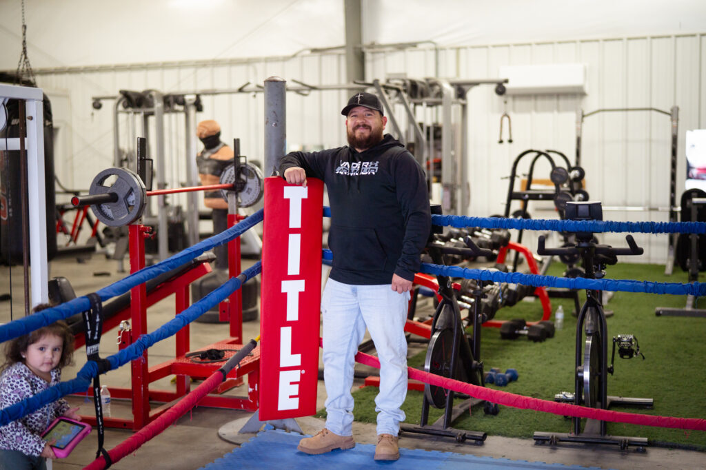A man wearing a black hoodie standing inside a boxing ring smiling while holding a red rectangular sign that reads "Title"