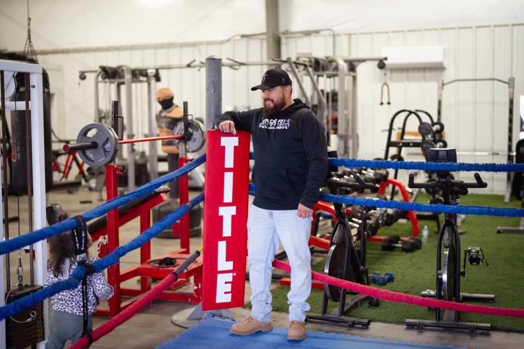 A man wearing a black hoodie standing inside a boxing ring holding a red rectangular sign that reads "Title"