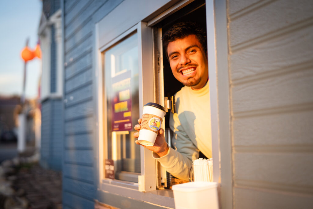 A drive-thru employee at a cafe smiling at the camera while holding a coffee cup advertising the business