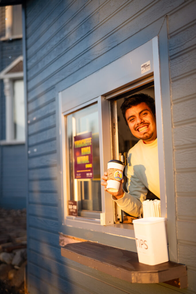 A drive-thru employee at a cafe smiling at the camera while holding a coffee cup advertising the business