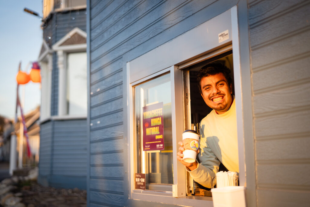 A drive-thru  employee at a cafe smiling at the camera while holding a coffee cup advertising the business