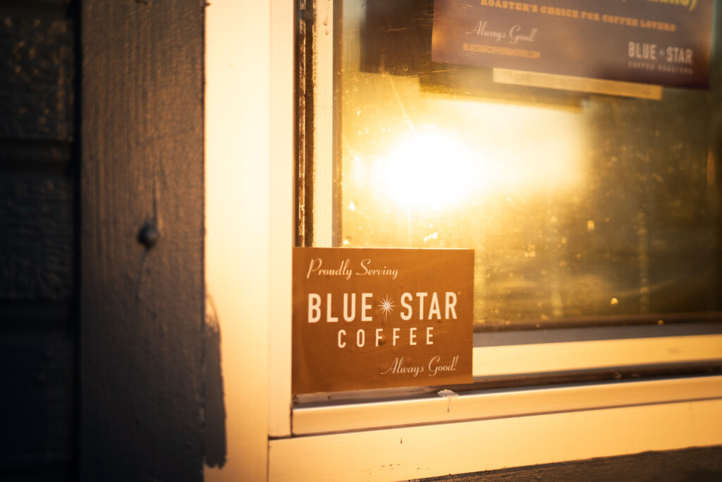 A sticker on a window lit by the sun that reads "Proudly Serving Blue Star Coffee, Always Good"