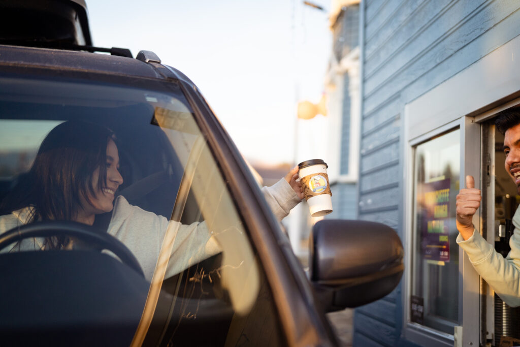 A woman smiling as she receives her coffee through the drive through window from a smiling male employee