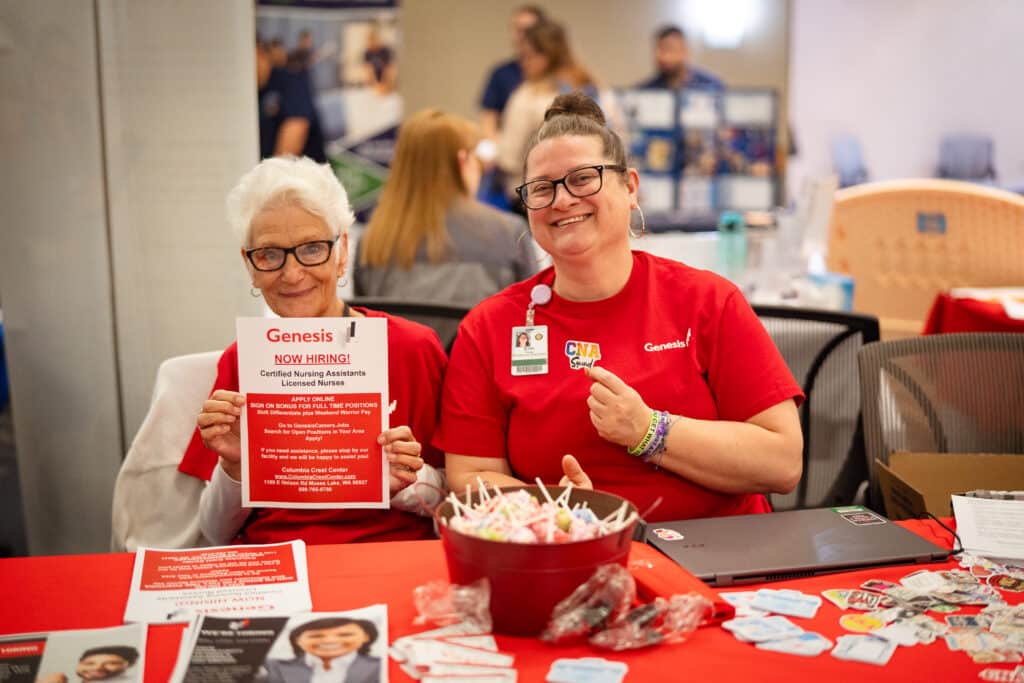 Two ladies at the BBCC Job Fair. Lady on the left with white short hair holding a "Now Hiring" flyer. To her right is a lady with brown hair in a bun holding a "CNA squad" sticker.