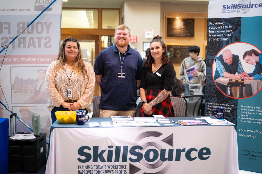 A group of 3 adults with big smiles standing behind a SkillSource table at the job fair at Big Bend Community College.