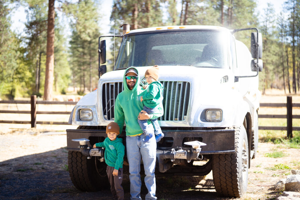 A man wearing a green hoodie standing in front of a white utility truck in a sunny day holding a toddler on one arm while a second child stands besides him