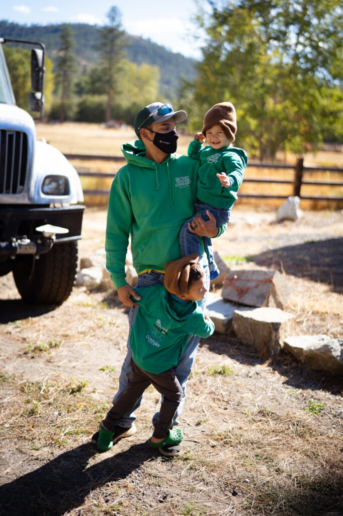 A man wearing a green hoodie and a black face mask standing outside in a sunny day holding a smiling toddler on one arm while a second child hugs his leg