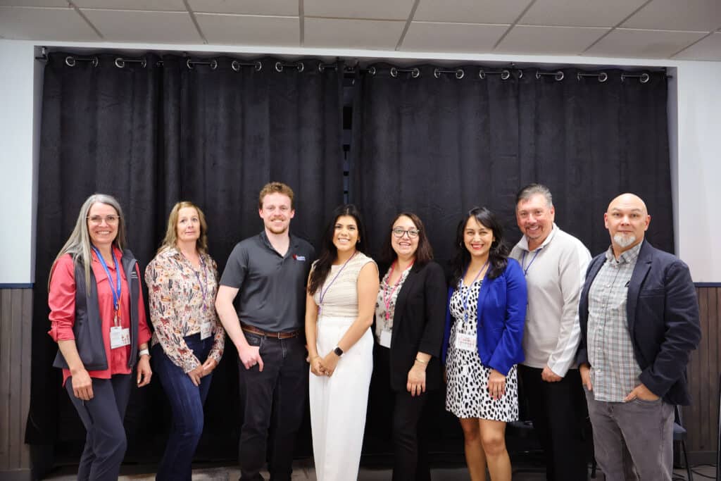 A group of eight smiling professional adults against a black curtain backdrop at a workforce collaboration summit.