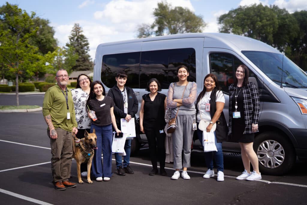 A group of eight people plus a German Shepherd service dog smile outside of a silver van.