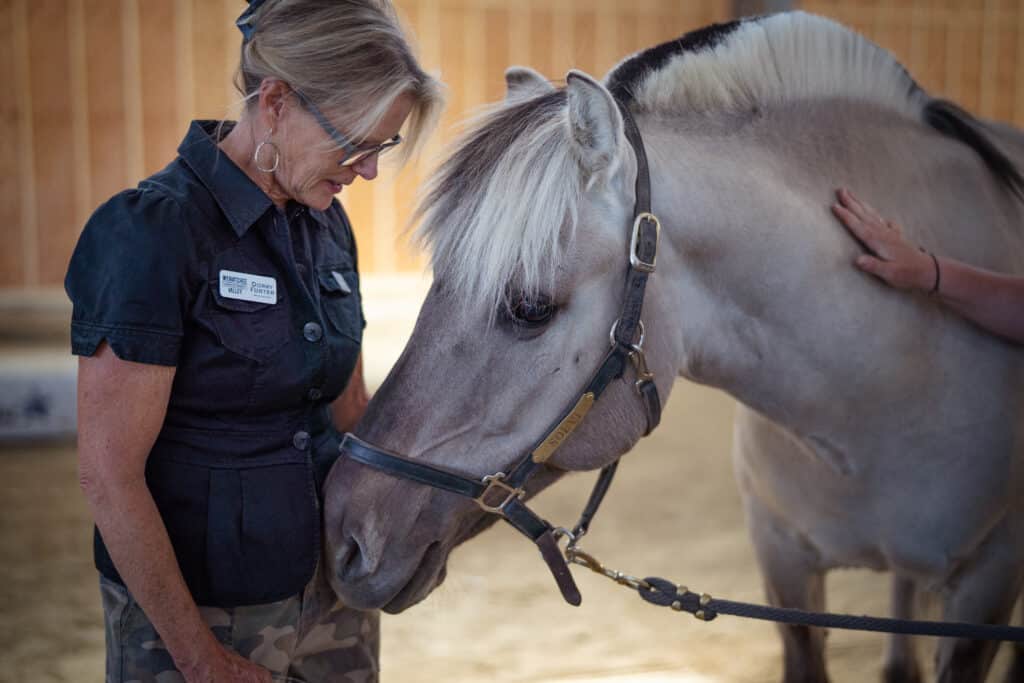 A woman in a dark uniform gently interacting with a light grey horse indoors