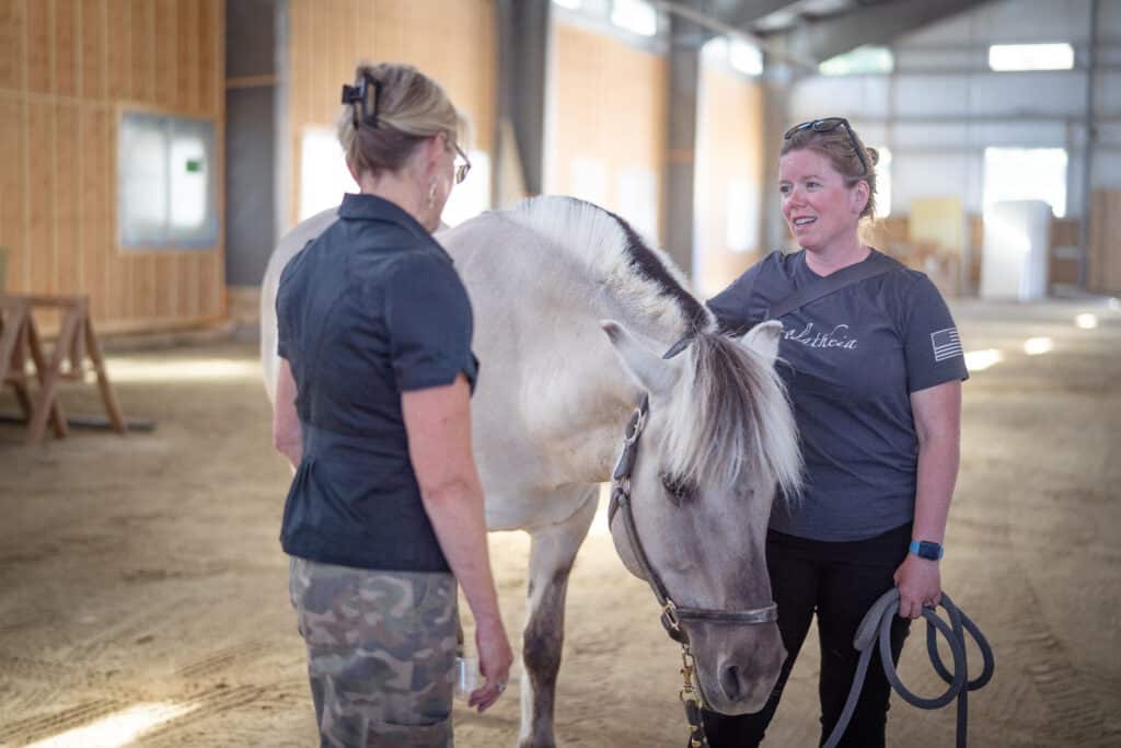 Two woman indoors smiling while having a conversation with a light grey horse in between them