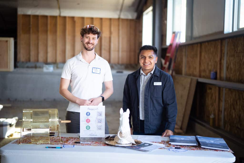 Two men smiling at the camera standing behind a table with flyers, man on the left is wearing a white collared shirt while man on the right is wearing a navy jacket