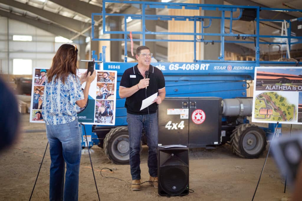 A man wearing a black top and jeans holding a mic, laughing while doing a presentation
