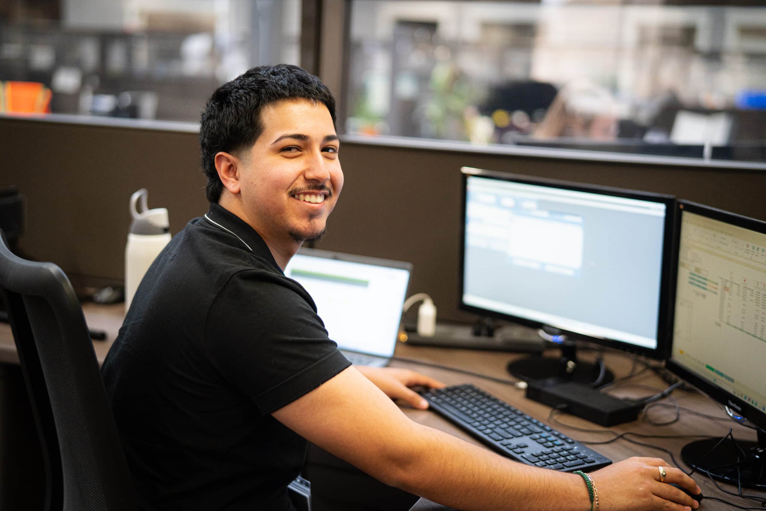 A smiling man sits behind a computer screen in a well lit office space.