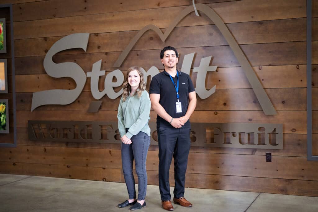 A woman and a man posing for a photo in front of a brown wooden wall with Stemilt's logo.