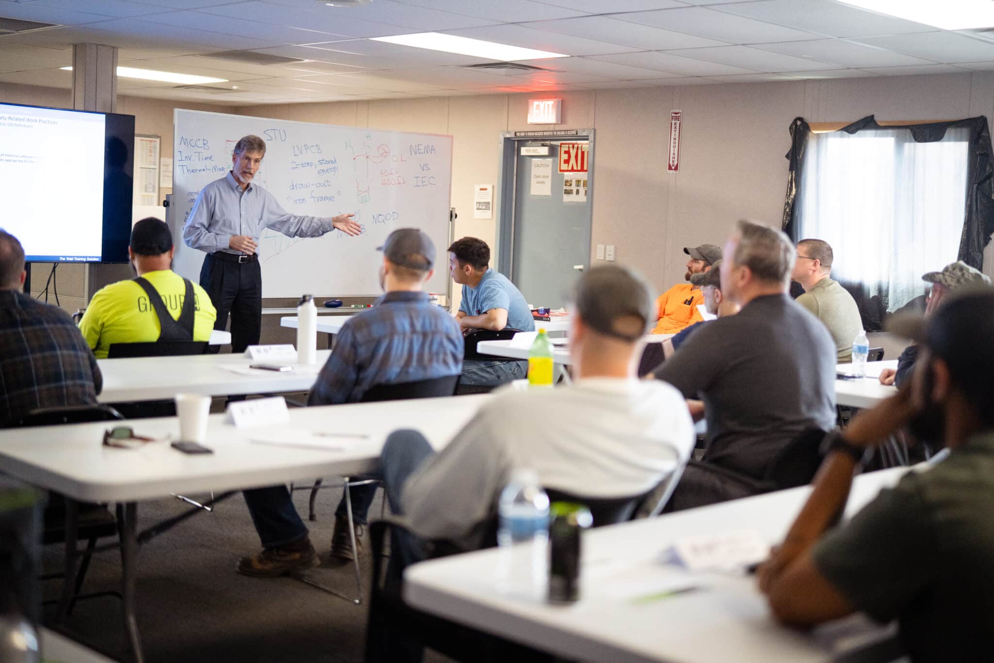 A man in professional attire stands at the front of a classroom setting before a group of adult workers who are seated at tables.