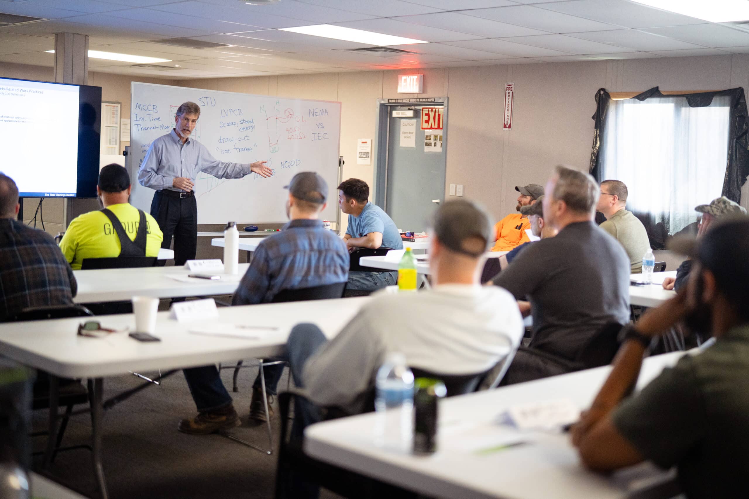A man in professional attire stands at the front of a classroom setting before a group of adult workers who are seated at tables.