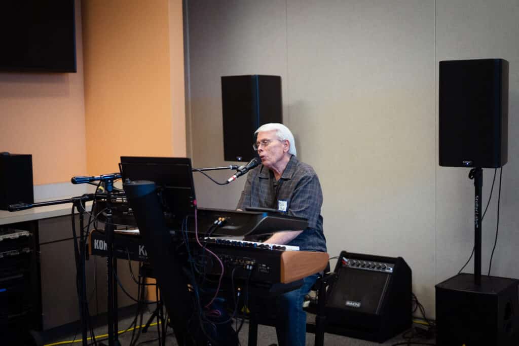 7-1-25 ML BAH-01 A man with a dark grey shirt and white hair is sitting down on a bench inside a music room singing and playing the piano