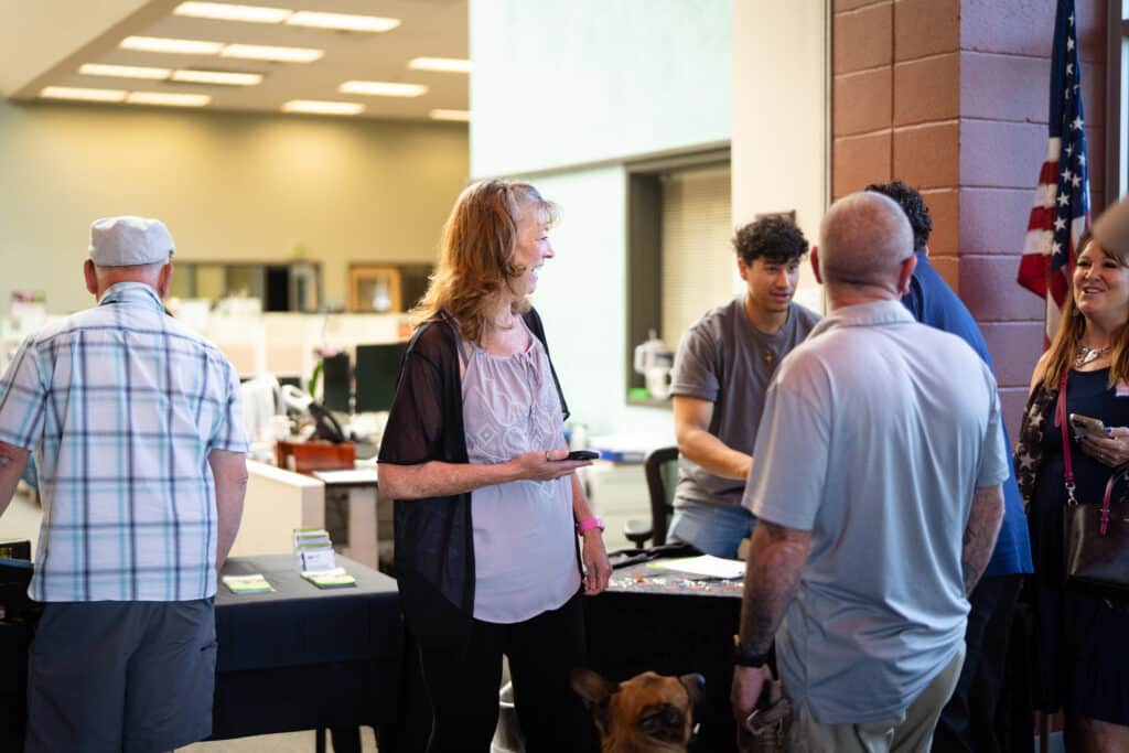 7-1-25 ML BAH-04 A group of people chat and engage at an indoor event with informational tables and a dog visible in the scene.