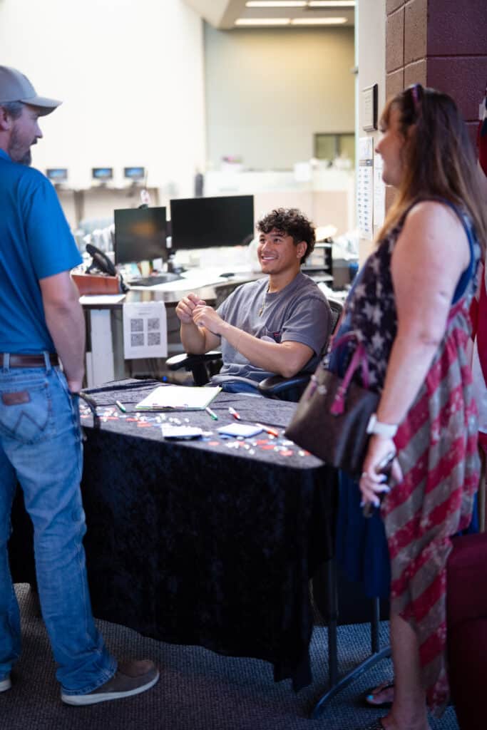 7-1-25 ML BAH-07 Two adults are standing in front of a informational table while talking to a young male sitting behind the table.