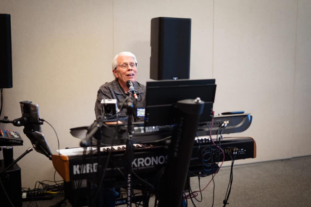 7-1-25 ML BAH-09 A male with white hair is sitting on a chair inside a well lit room while singing and playing the piano.