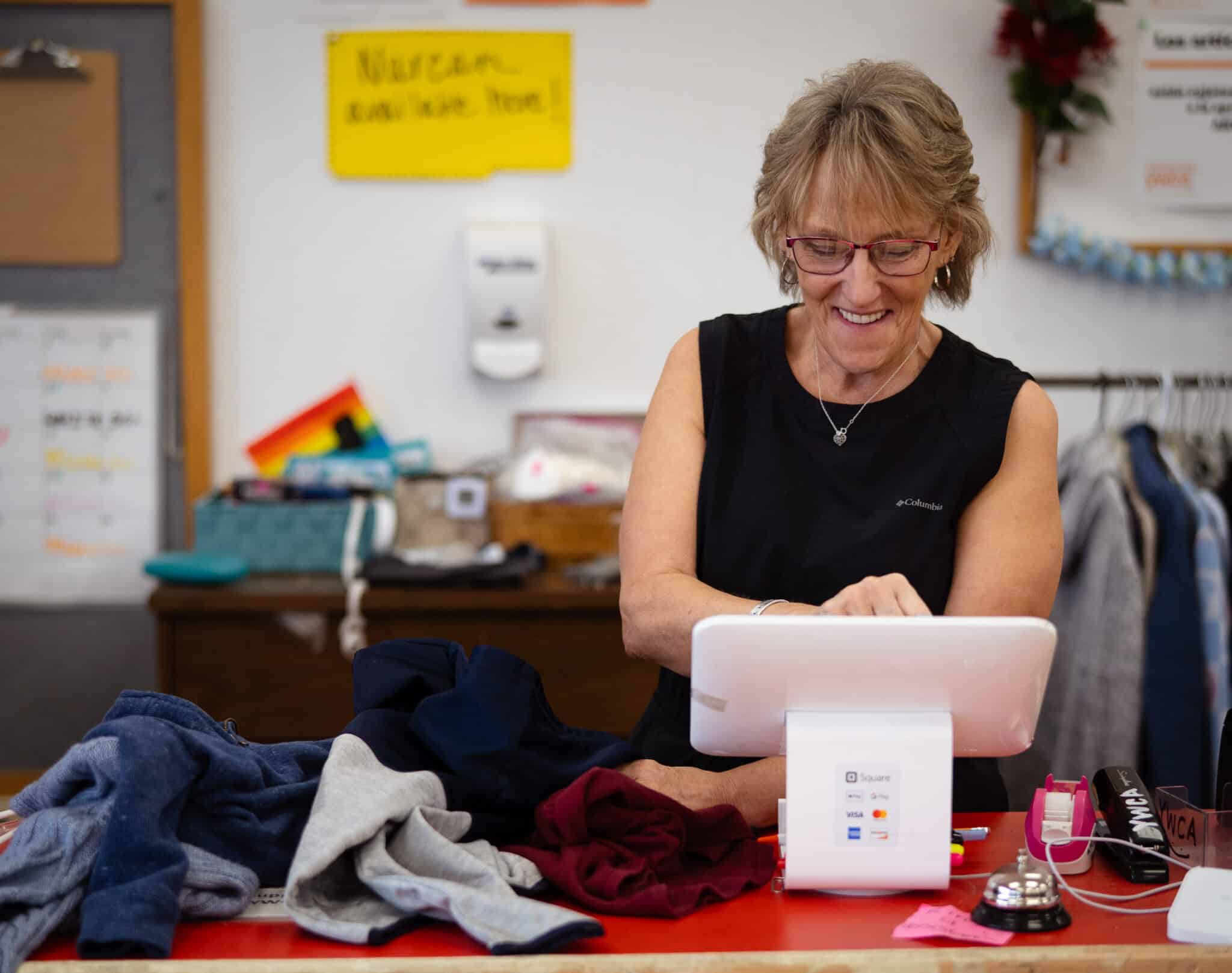 Woman wearing a black top is smiling behind a cash register.