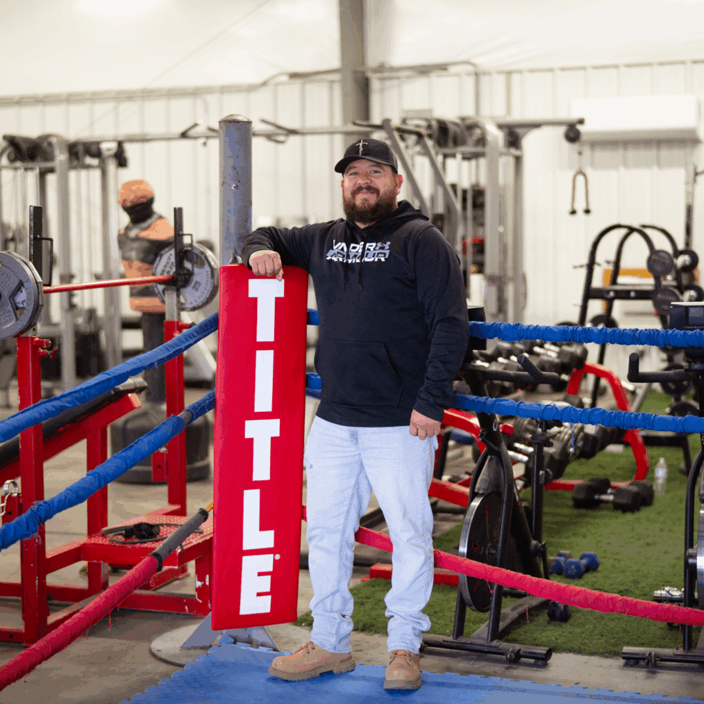 A boxing gym owner leans against the padded red railing of a boxing ring.