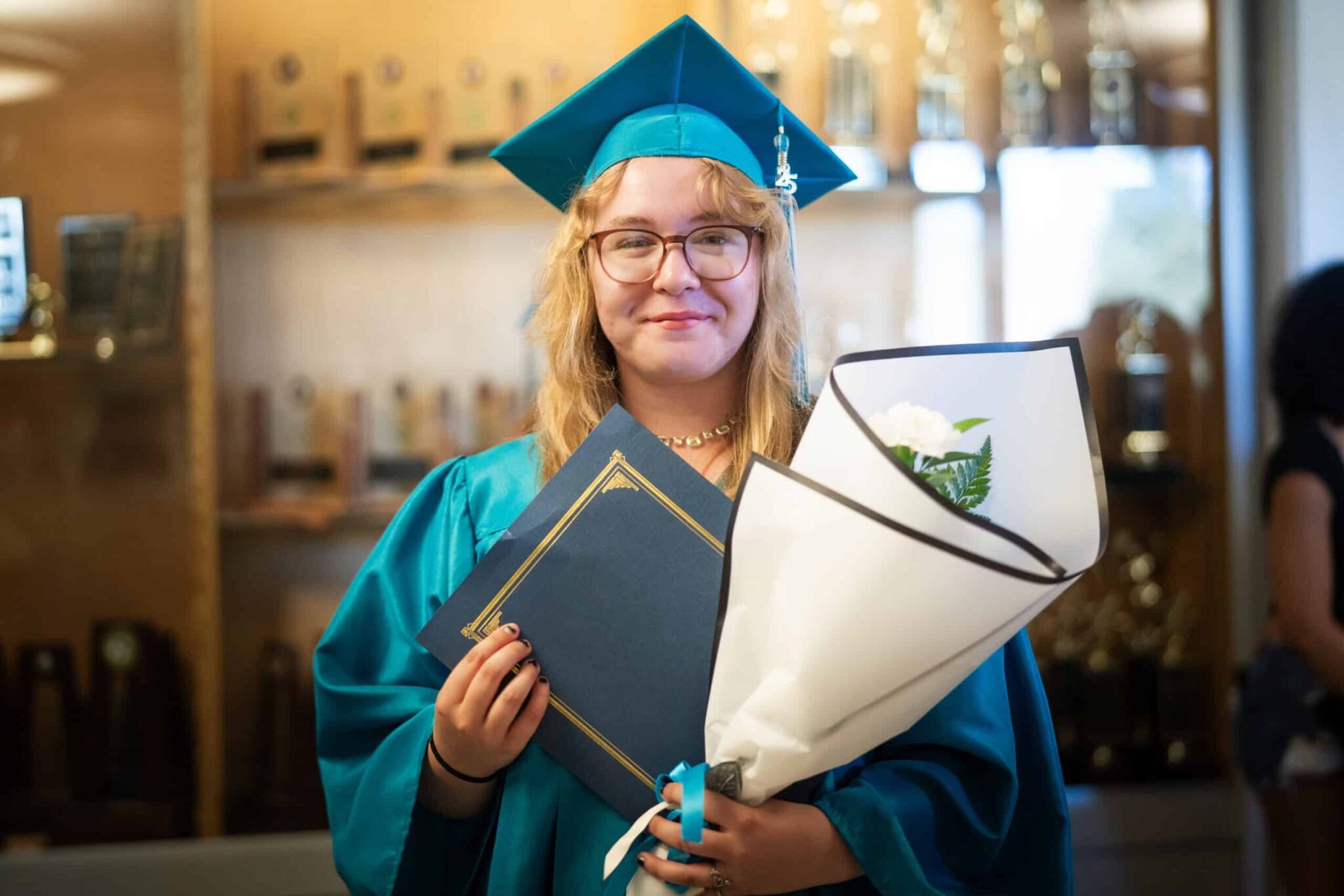 Graduate wearing a teal cap and gown smiling holding a diploma.