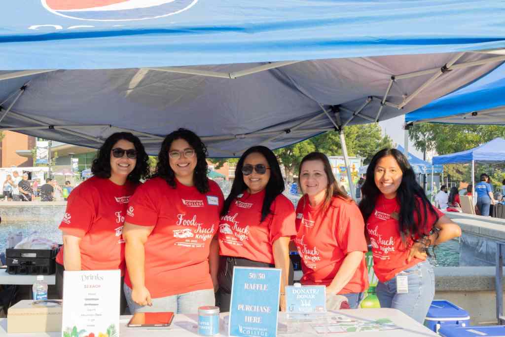5 brunette women are wearing red shirts and are smiling at a camera while standing behind a booth at Wenatchee Valley College.