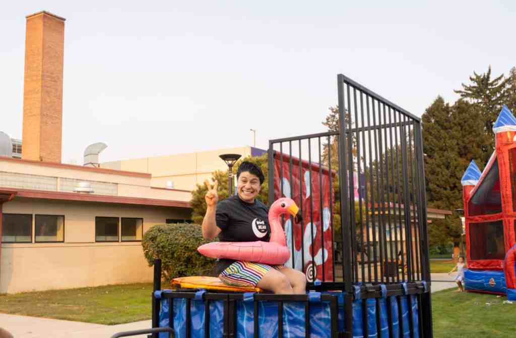 A woman with short hair and grey t-shirt is smiling holding up a peace sign while sitting in a dunk tank with a flamingo floatie.