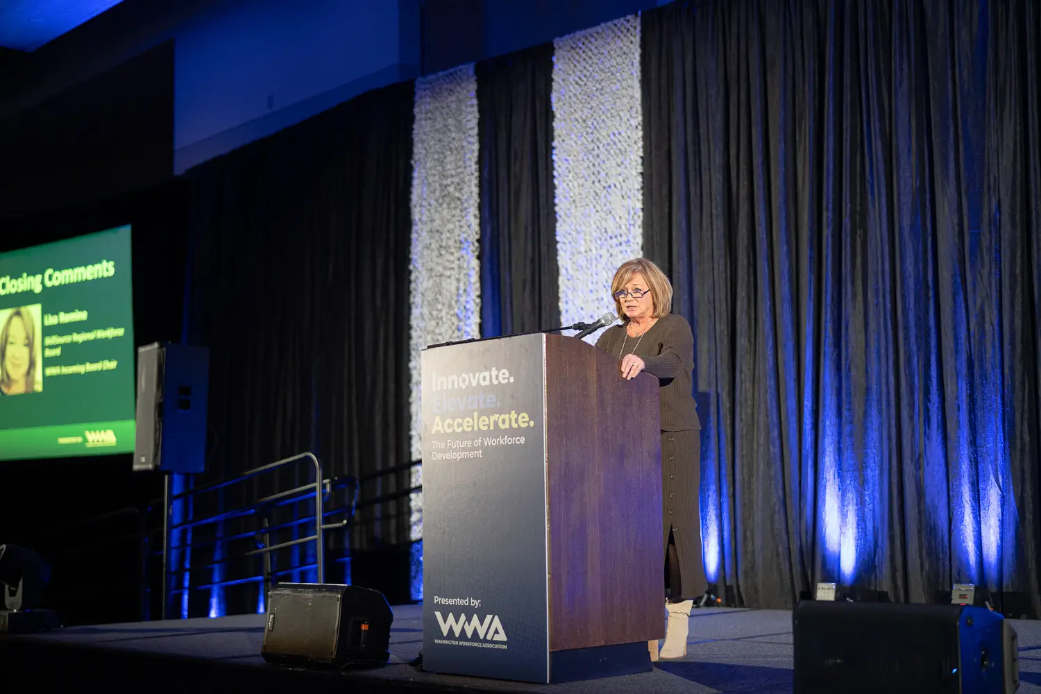 A woman in glasses and with short blonde hair stands on stage behind a podium against a blue backdrop.