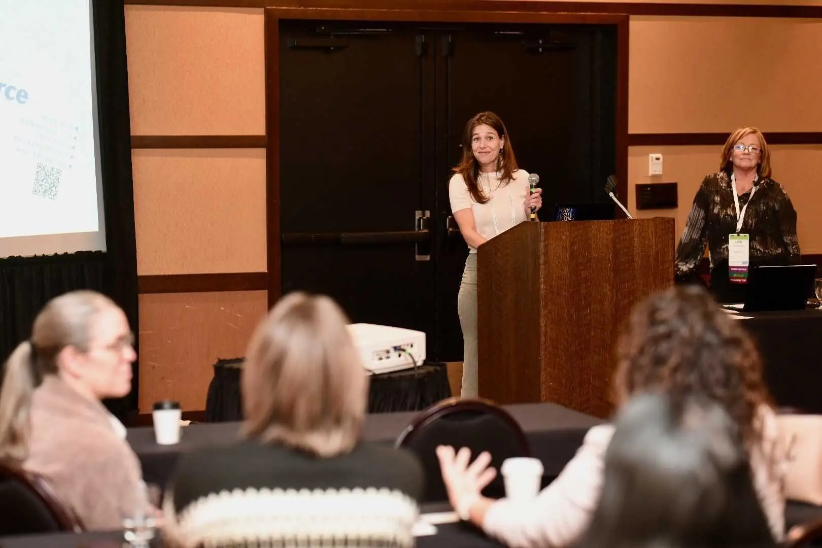 A long-haired female in a white blouse stands behind a podium at the front of conference room.