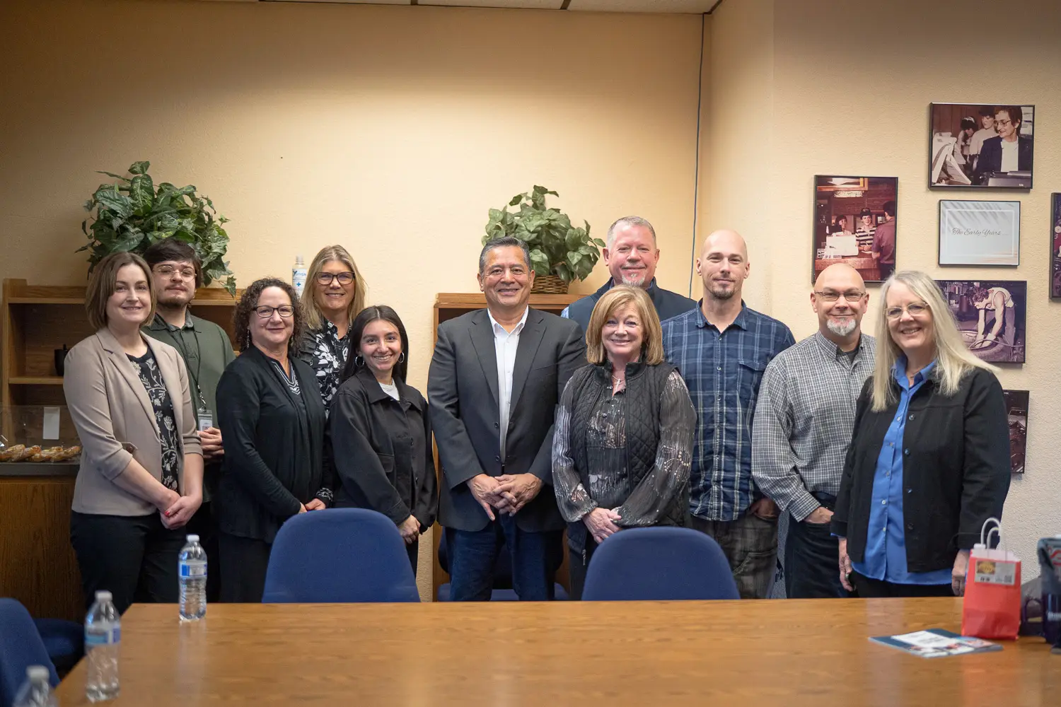 A group of diverse professionals stand behind a conference table with a local elected official at the center and everyone smiles in the direction of the camera.