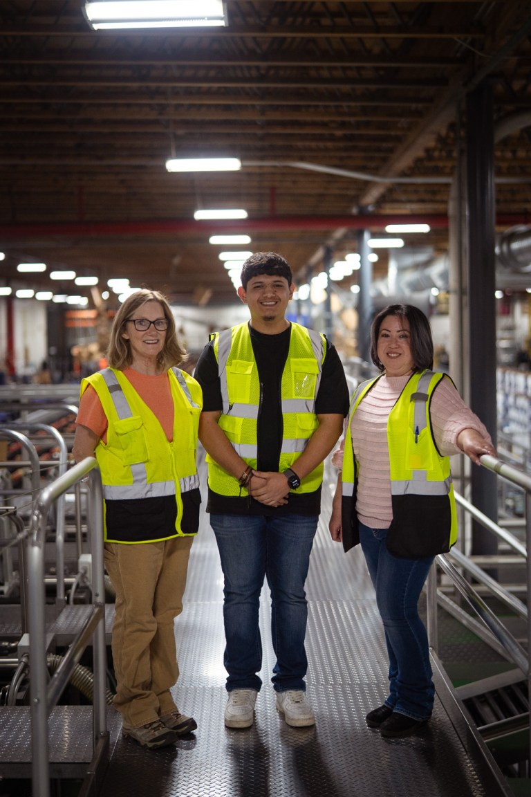 Two women and a young man wear neon yellow safety vests on a metal walkway inside a warehouse.