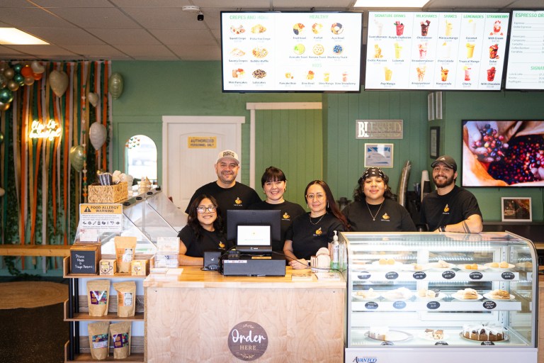 Six people in black t-shirts smile while standing together behind the cafe counter where they all work.