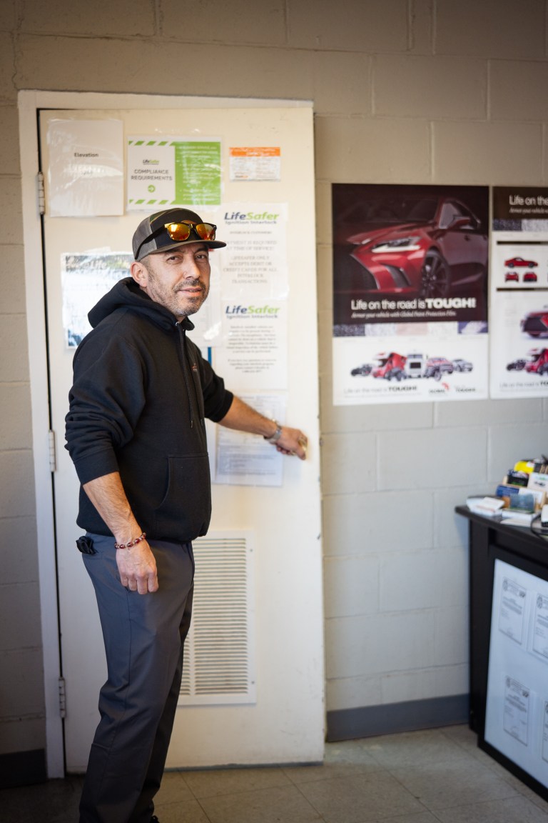 A man in a black jacket and wearing a cap with sunglasses on the brim has his hand opens the shop door inside his auto tint business.