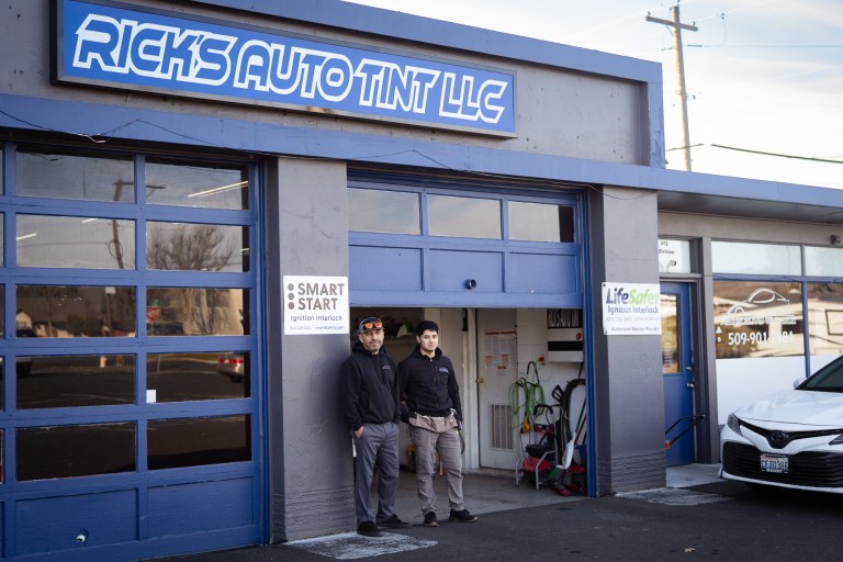 Two men stand outside of a garage door beneath a sign that says Rick's Auto Tint LLC