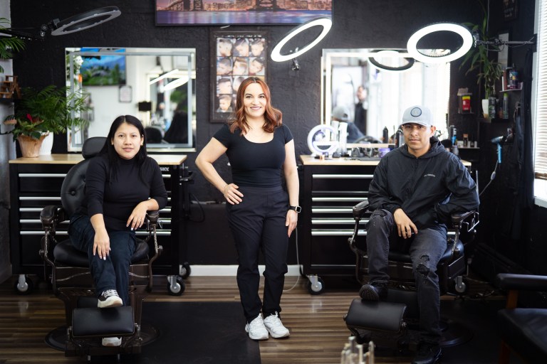 Three stylists all wearing black pose near barber chairs in a salon.