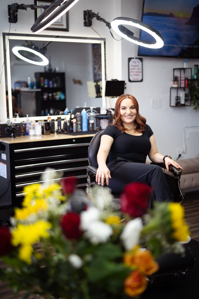A woman wearing all black sits cross legged in a salon chair with a bouquet of yellow and red flowers in the foreground.