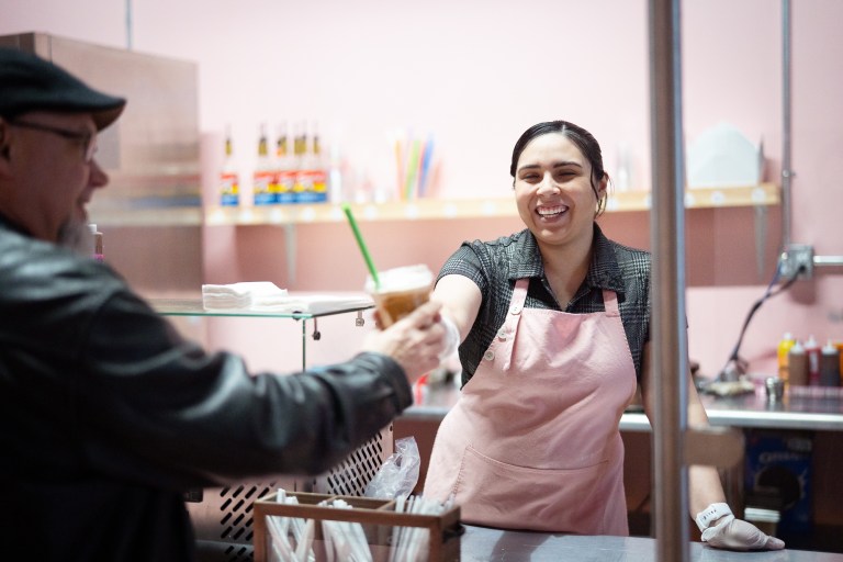 A smiling woman wearing a pink apron reaches across a shop counter to hand a customer a milkshake.