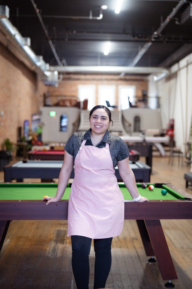 A latina business owner wearing a light pink apron stands in front of a green topped pool table.