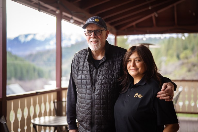 An older gentleman in in glasses and a ball cap stands with his arm around the shoulder of his dark-haired wife on the front porch of their wine business with mountains in the background.