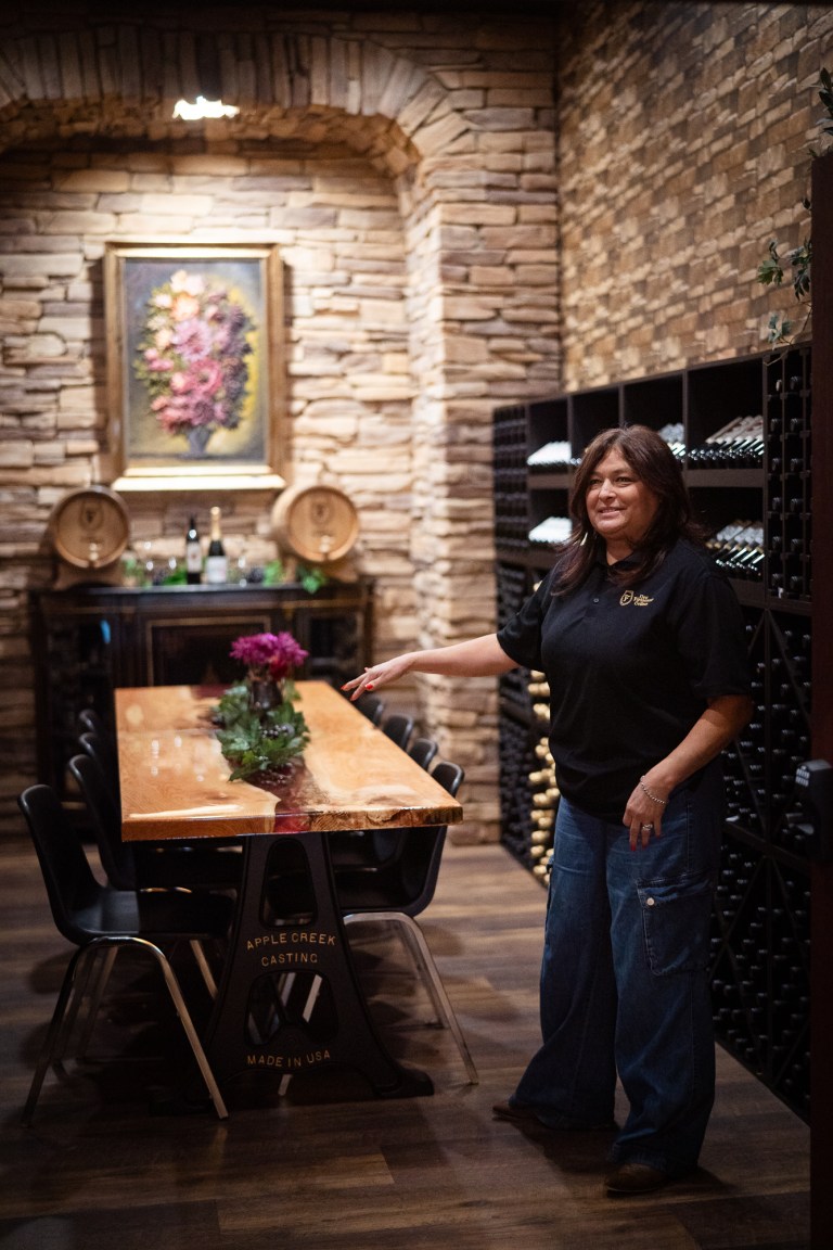 Inside a wine tasting room with a handcrafted wooden table in the middle and a dark haired woman stands near gesturing to a wine-colored inset that runs the length of the table.